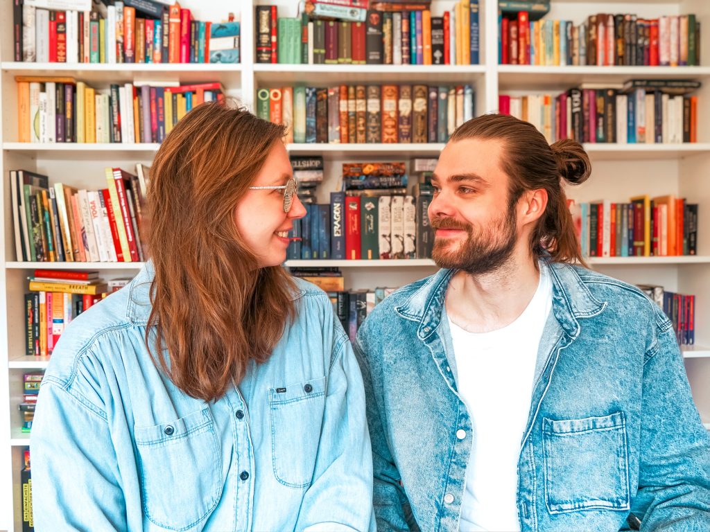 Jukes Studio Teamfoto, Rike und Julius gucken sich an und sitzen vor einem Bücherregal, Webdesign und Online Marketing für dein Business