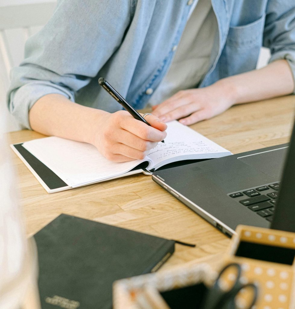 Adult writing notes on a notebook while working on a laptop in a cozy home office environment.