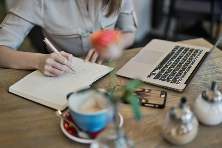 Frau schreibt in Notizbuch zwischen Laptop und Kaffeetasse zum Thema Website-Texte